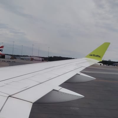 The image captures a view from an aircraft window on the ground at Stockholm-Arlanda Airport in Sweden, under an overcast sky. The prominent subject is the right wing and bright green tail fin of an AirBaltic aircraft, seen from inside the cabin. The tail displays the "airBaltic" logo in black lettering. The wing is white with visible panel lines and control surfaces (flaps/ailerons).
On the tarmac to the left of the wing, a portion of another aircraft's tail fin is visible, featuring red, white, and blue markings (consistent with British Airways). Further in the background, a series of low-rise airport buildings or hangars are present, with several tall light poles erected on the apron. Yellow ground support equipment and luggage carts are visible near the buildings and on the tarmac. A distant treeline can be seen beyond the airport infrastructure, under the gray, cloudy sky. The tarmac itself is dark gray with distinct expansion joints and markings.