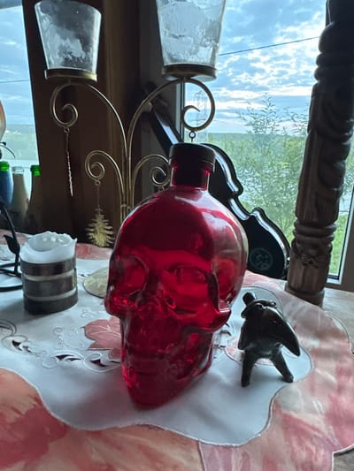 The image shows a tabletop with various objects placed in front of a window. A large, red, skull-shaped glass bottle is prominently displayed in the center. To the left of the bottle is a white candle in a wooden holder. Behind the candle, part of a gold candelabra with a hanging ornament is visible. To the right of the skull bottle, a small, dark figurine of a creature with a long snout and prominent eyes stands on the table. The background shows a window with a view of trees and a cloudy sky. A wooden carved pillar is partially visible on the right side of the image. The objects are arranged on a tablecloth with a floral pattern. There are no people or actions depicted in the image. The scene appears to be indoors, near a window, in what could be a domestic setting. No specific details are provided to place the scene in Sergeevka, Russia.