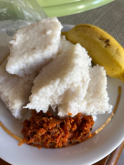 A white plate containing a portion of cooked white rice molded into rectangular shapes, a small pile of orange-red relish, and a ripe yellow banana is displayed on a table with a patterned surface. A portion of a green object and a translucent plastic bag are visible in the background, suggesting an indoor setting, possibly in Dehiwala, Sri Lanka, where such a meal might be served.