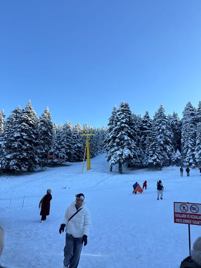 The image depicts a snowy mountain scene, identified as a ski resort, likely Uludağ, near Bursa, Turkey. The environment is characterized by a clear, bright blue sky above and extensive snow cover across the ground and trees.

In the mid-ground, a prominent yellow metal ski lift tower stands, supporting a cable from which red chairlift chairs are suspended and moving uphill. The slopes are bordered by dense clusters of snow-laden evergreen trees, forming a thick forest.

Approximately ten individuals are visible on the snowy slope. In the foreground, a male subject wearing a white puffer jacket, blue jeans, black gloves, and white earmuffs stands, looking slightly to the right with a smile. A black strap from a bag runs across his chest. To his left, another individual in a long maroon coat and light-colored head covering stands with their back partially to the viewer.

Further into the mid-ground, two individuals, one in a red jacket and another in a dark jacket, are observed near a red sled-like object on the snow. Other individuals are scattered across the slope in the mid-ground and background, wearing various winter clothing, some standing, others seemingly engaged in recreational activities.

In the lower right foreground, a red and white warning sign is visible. The Turkish text on the sign reads "YAYALARIN VE KIZAKLARIN PİSTE GİRMESİ YASAKTIR," which translates to "PEDESTRIANS AND SLEDDERS ARE NOT ALLOWED ON THE PISTE." The sign includes pictograms depicting a person walking and a person sledding, both with a prohibitory slash.