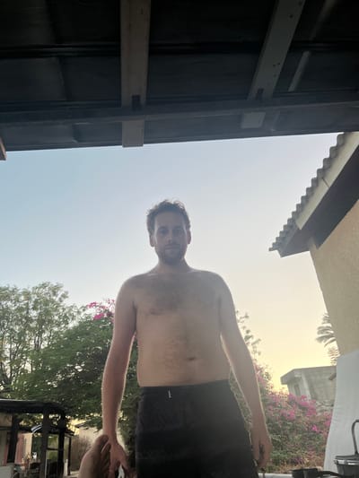 An adult male stands outdoors, bare-chested and wearing dark swim shorts, looking directly at the camera. He has short brown hair and a light beard. His left foot is partially visible in the lower-left foreground. Above him is the dark underside of a covered structure with exposed wooden beams. The background features a clear sky transitioning from light blue to a yellowish-orange hue, suggesting late afternoon or early morning light. To the left, green trees with pink blossoms are visible. To the right, a building with a tiled roof and a palm tree are present, along with more pink flowering bushes and a distant light-colored building with a flat roof. The scene is located in Emek HaMaayanot Regional Council, Israel.