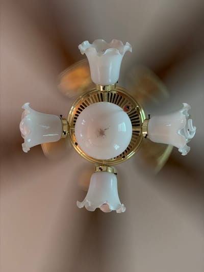 This is a close-up, overhead shot of a brass ceiling fan with four white, tulip-shaped glass light fixtures. The fan blades are in motion, creating a slight blur that gives the image a dynamic feel. The central part of the fan is illuminated by a spherical white light bulb, and the brass accents of the fan's motor housing are visible. The light fixtures have decorative, wavy edges. The background is a soft, blurred beige, likely a ceiling. The overall impression is one of an active, functional object in a domestic setting, likely indoors. There is no visible text in the image. The time of day and weather are not determinable from the image. The location context provided is Stallings, United States of America.