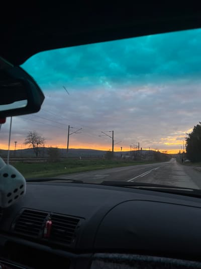 The image, taken from inside a car, shows a sunset over the railway tracks and a road in Dalgopol, Bulgaria. The road runs parallel to the railway, and both lead towards the horizon where the sun is setting. Overhead power lines are visible alongside the tracks. The sky is a mix of blues, grays, and oranges, indicating the time of day. Car interior elements, such as the dashboard, air vents, and a rearview mirror, are visible in the foreground, giving the viewpoint.