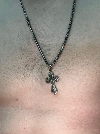 A silver cross necklace is centered on a person's chest, which is covered in fine, light-colored hair. The necklace chain is made of interlocking links and is slightly twisted near the top, where it appears to have a clasp or adjustment mechanism. The cross itself has a detailed design, with a raised figure at its center. The skin appears pale with some minor variations in tone and the presence of a few small moles. The background is out of focus, suggesting a close-up shot. The scene takes place in Corlăteni, Moldova.