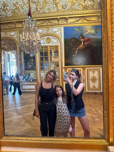 A golden framed mirror reflects a richly decorated room, presumably a palace, featuring ornate molding, paintings, a chandelier, and parquet flooring. Three people are visible in the mirror's reflection. An adult in black stands in front, with two younger people behind them. The person closest to the top is holding a phone and seems to be taking a picture. Other visitors are also visible in the reflection below.