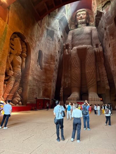 The image depicts the Leshan Giant Buddha, a massive stone sculpture in Shizhong, China. The Buddha is carved into a cliff face, with its head and torso visible in the top left portion of the image. The legs extend downwards, towards a walkway where a group of people are standing. The people appear to be tourists, observing the Buddha. A second stone carving is visible below the walkway. The environment appears to be a temple or historic site, with a reddish-brown color palette due to the sandstone material.