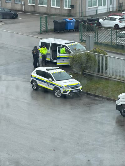 The image presents an outdoor scene depicting police activity in Logatec, Slovenia, under overcast or post-precipitation conditions, evidenced by the wet asphalt pavement.

In the foreground, a white Slovenian police SUV, identified by "POLICIJA" markings and blue and yellow stripes, is parked facing right. Its front license plate, "P*06-889", is visible. Behind and slightly to the left of the SUV, a white Slovenian police van, also with "POLICIJA" markings and blue and yellow stripes, is parked. The driver's side sliding door of the van is open. Both police vehicles are equipped with rooftop light bars.

Three individuals are present near the open police van. Two individuals in uniform are standing on the pavement. One individual wears a dark, tactical-style uniform with a vest and is positioned to the left, facing towards the open van. Another individual, wearing a high-visibility yellow police jacket with reflective stripes and dark uniform pants, stands between the first individual and the van, facing the individual in the dark uniform. A third individual, also in a high-visibility yellow jacket, is partially visible inside the open police van.

The background features a light-colored commercial or industrial building with multiple windows. A green metal fence runs horizontally across the midground, separating the primary paved area from a grassy strip and an additional paved parking area. Behind this fence, several large black and blue industrial waste bins are visible. A dark-colored sedan is parked on the far left in the background, and a white hatchback is parked on the far right. A portion of a third white vehicle is visible in the extreme right foreground.