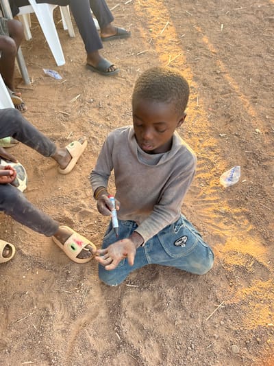 The scene is set outdoors in Barakin Acha, Nigeria, on reddish-brown, dusty ground with scattered small stones, dry vegetation, and white plastic debris. A pre-adolescent male with short, dark, textured hair kneels on his right knee in the foreground, focusing intently on his left open palm. He wears a worn, long-sleeved grey top with a cowl neckline and worn blue denim jeans featuring a distinct "White-Oil" design on the right thigh pocket. Multiple thin, yellow wristbands are visible on his left wrist. His left palm shows a red liquid substance, while his right hand holds a cylindrical, white marker-style object with a red tip. In the background and to the left, partial views of other individuals' limbs are present, some seated on white plastic chairs. Visible footwear includes black slides, tan slides, and tan slippers with prominent eye-like decorations. A white and black soccer ball is partially visible in the lower-left quadrant. Natural daylight illuminates the scene, characterized by warm tones consistent with late afternoon.