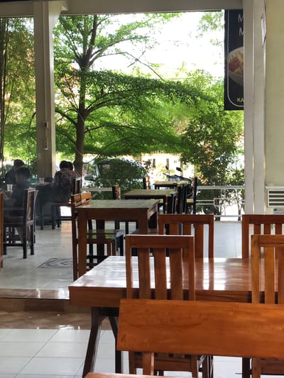 The image captures an outdoor seating area of a restaurant or cafe, likely in Keudealue Ie Puteh, Indonesia, characterized by its open-air design with a roofed structure. The scene is dominated by wooden tables and chairs, arranged on a tiled floor. Lush green trees and foliage are visible through the open space, creating a natural, serene backdrop. Several patrons are seated at tables, engaged in conversations or meals. The lighting suggests it is daytime, possibly late morning or afternoon, with natural light filtering through the leaves. A sign is partially visible on the right, featuring text and an image of food. The overall ambiance is relaxed and inviting, with a blend of indoor seating and outdoor natural elements.
