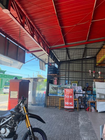 A motorcycle is parked in the foreground of an outdoor marketplace. The motorcycle is black with gold colored front suspension. Behind the motorcycle, to the left, is a red cabinet. To the right of the cabinet, there are stacks of bottled water and shelves. A metal frame structure supports a red roof overhead. In the background, a green building with a white patterned facade is visible. The ground is paved with brick. There are no people visible in the image.