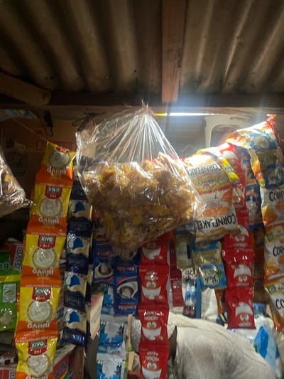 This image depicts a small shop in Sauka, Nigeria, likely a kiosk or a roadside stall. The setting is cramped, with shelves overflowing with various packaged goods, predominantly snacks and food items. The items are stacked high, creating a colorful and somewhat chaotic display.
Prominently visible are bags of "Cornflakes," "Happy Chinchin," and "Garri," which is a staple West African food. There are also packets of "Kivo" and "Topcafe," suggesting a mix of food and beverage products. The packaging displays bright colors and imagery, including cartoon characters on some of the "Chinchin" bags. The goods are arranged in rows, with some items, particularly a large bag of fried snacks, appearing to be loose in a plastic bag in the foreground.
The lighting suggests it might be daytime, with sunlight streaming in from the left, illuminating the products and casting some shadows. The wall to the left appears to be made of corrugated metal, a common building material for such small businesses in the region. The overall impression is one of abundance and everyday commerce in a local Nigerian setting. The image captures the vibrant nature of small-scale retail operations, offering a glimpse into the available consumer goods.