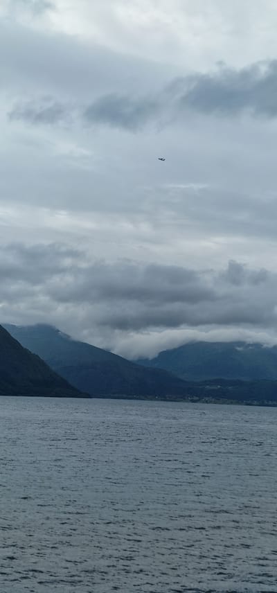 An airplane is visible in the sky above a body of water and mountains. The sky is overcast with gray clouds. The body of water is dark and appears to be a fjord or lake. The mountains are covered in dark green vegetation, and some of the peaks are obscured by clouds. The scene is likely taken from a boat or the shore. The image appears to be a landscape shot taken on a cloudy day in the vicinity of Volda, Norway. There are no people visible in the image.