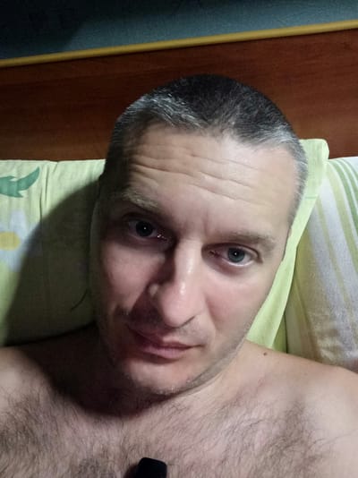 A close-up, selfie-style photograph shows a light-skinned adult male with short, graying hair and visible chest hair. He is lying down, propped up on a pillow with a green and white striped pattern and a floral design on the left side. The background consists of a dark blue surface above a brown wooden headboard. The image is illuminated by soft light, creating shadows on his face and chest. There is no indication of the city of Balakliya, Ukraine, or any other specific location within the frame. The man's expression is neutral. A small, dark object, possibly a phone, is visible near his collarbone.