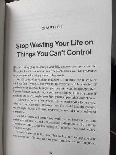 The image displays an open book, captured at a slight angle, showing the right-hand page with printed text. The scene is located in Breda, Netherlands. The page is white with black print. "CHAPTER 1" is centered near the top of the page. Below this, the chapter title, "Stop Wasting Your Life on Things You Can't Control," is presented in a larger, bold typeface, spanning two lines. Body text follows, beginning with a dropped capital 'I', discussing themes of personal control and external validation. A visible excerpt reads: "If you're struggling to change your life, achieve your goals, or feel happier, I want you to hear this: *The problem isn't you. The problem is the power you unknowingly give to other people.*" Further paragraphs continue this discussion. The page number "17" is centered at the bottom of the page. The book's left binding is visible, and the background is blurred, consistent with an indoor setting. No individuals are present.