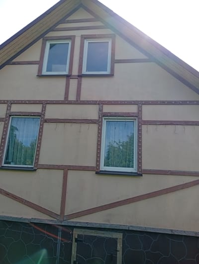 A close-up, eye-level, exterior shot captures the upper half of a light beige-colored house in Lavriki, Russia. The house features a prominent gable roof with wooden trim and two rectangular windows positioned side-by-side on the upper level. Below these, on the main facade, are three more rectangular windows, also with white frames and sheer white curtains. Decorative brown wooden beams, in a Tudor-style pattern, are affixed to the beige siding, creating a grid-like effect. A string of small, faceted lights, reminiscent of Christmas lights, is draped horizontally above the lower row of windows and vertically around their frames. The lower portion of the house visible in the frame has a dark gray siding with a light gray, abstract pattern resembling cracked earth or stylized scales. A small wooden structure with a dark-covered opening is visible on the far right. The overall impression is one of a residential building, possibly undergoing or having recently undergone decorative enhancements.