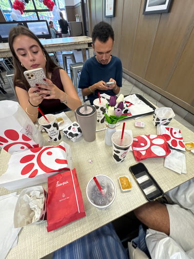 The image depicts a table laden with Chick-fil-A food and beverages, suggesting a casual dining experience at a restaurant. Two individuals, a woman and a man, are seated at the table, both engrossed in their smartphones, ignoring their meals and drinks. The woman, on the left, is holding her phone and appears to be typing. The man, on the right, is also looking at his phone. The table is covered with various Chick-fil-A branded items including several white paper bags with red logos, red and white cow-print boxes, cups with straws, and a small packet of sauce. In the background, there are balloons, suggesting a celebratory atmosphere, possibly a birthday or special event. The setting appears to be an indoor dining area, with wooden paneling on the walls and metal stools visible. The presence of the balloons and the arrangement of the food indicate a possibly social gathering, though the focus on phones suggests a disconnect among the diners. The woman's phone case is a white patterned design, and she is wearing rings and bracelets. The man has a silver bracelet on his wrist. The overall scene conveys a modern dining scenario where technology often takes precedence over in-person interaction.