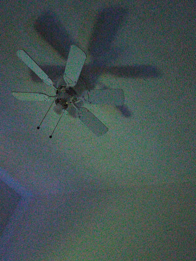 The image shows a ceiling fan with four blades, viewed from below. The fan appears to be made of a white material and is mounted on the ceiling. Two dark pull cords hang down from the fan, each with a dark spherical knob at the end. The ceiling is textured and appears to be painted a light color, casting a shadow from the fan. The lighting in the room is dim, and the image has a significant amount of digital noise. There is a faint blueish light source visible in the lower left portion of the image, which may be a reflection or an external light source. The overall scene is static and does not depict any activity. The location is not discernible from the image itself.