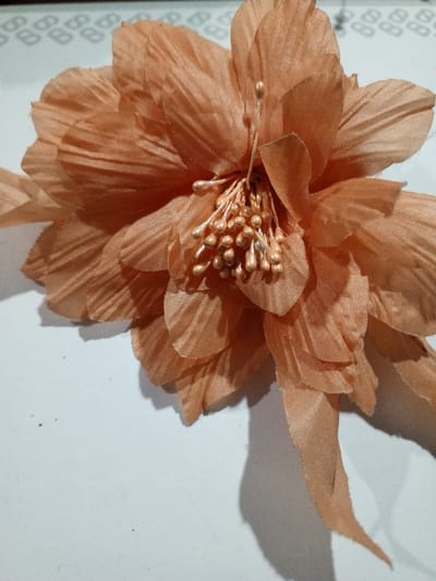 A peach-colored fabric flower with multiple layers of petals sits on a white surface. The petals have a subtle ribbed texture. In the flower's center are numerous stamens with bead-like tips, some metallic gold and some slightly tarnished silver. A few loose strips of fabric extend from the base of the flower. The overall presentation suggests a crafted or decorative item.