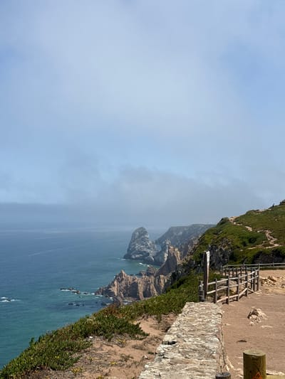 This is a landscape photograph taken at Cabo da Roca, the westernmost point of mainland Europe, near Colares, Portugal. The image captures a dramatic coastal view with rugged cliffs meeting a vast expanse of deep blue ocean. In the foreground, a sandy path runs alongside a rustic wooden fence and a low stone wall. Vegetation, including green succulents and small yellow flowers, grows along the cliff edges. In the distance, several rocky outcrops emerge from the sea. The sky is partly obscured by a light haze or fog, creating a soft, diffused light. No people are clearly visible in the frame, although a distant figure can be seen on the hillside on the right. The overall scene conveys a sense of natural beauty and the raw power of the coastline.