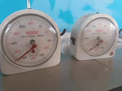 The image displays two vintage-style mechanical stopwatches or timers, made of white or off-white plastic or metal casing, resting on a flat, light grey-beige surface. The background is a blurry light blue wall with lighter, cloud-like or abstract patterns.

The stopwatch in the foreground is in sharp focus and is positioned slightly to the left of the frame's center. It features a round white face with clear black and red markings. The main dial indicates seconds, marked by black numbers at increments of 5 (5, 10, 15, 20, 25, 30, 35, 40, 45, 50, 55, 60), with smaller tick marks for individual seconds. The numbers 5, 10, 15, 20, 25, 30, 35, 40, 45, 50, 55, and 60 are printed in red. A large, prominent red second hand points to approximately 35 seconds. In the upper central part of the dial, the brand name "NDDC" is written in red capital letters, with "SECONDS TIMER" in black capital letters directly below it. There is a smaller, inner dial with a red hand and red markings, which appears to count minutes. This smaller red hand points slightly past the "30" mark on its own scale. The casing of this timer shows some signs of age or wear, with minor discoloration or marks. A metal loop or hook is visible at the top, and another metal component extends from the right side of the casing.

The second stopwatch is positioned further back and to the right, appearing slightly out of focus. It has a similar design to the front one, with a white casing and a visible dial. Although less clear, the "NDDC" and "SECONDS TIMER" labels are discernible on its face, along with the red numerical markings. Its large red second hand also appears to be pointing to a different time than the front timer. The casing of this timer also exhibits some wear.

Both timers appear to be stationary, suggesting they are either waiting to be used or are displayed. The overall impression is one of functional laboratory or sports timing equipment.