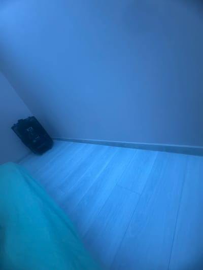 The image shows a corner of a room. The walls are light blue, and the floor is made of light-colored wooden planks. A black object, possibly a remote control or electronic device, lies in the corner where the wall meets the floor. A portion of a light green blanket or fabric is visible in the bottom right corner of the frame. The location is not discernible from the image.