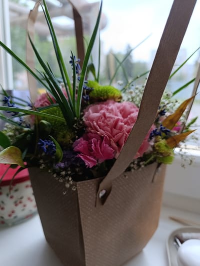 A close-up shot shows a bouquet of flowers in a brown paper bag. The bouquet contains several pink carnations, green pom-pom flowers, and small blue flowers, possibly lavender, interspersed with delicate white baby's breath. The bag has two brown paper handles that extend upwards. The flowers are arranged in a decorative manner within the bag, which is placed on a white surface, possibly a windowsill.
In the background, out of focus, there is a glimpse of a window with greenery and possibly buildings visible outside, suggesting an indoor setting. To the left of the bouquet, a portion of a bowl with a red cherry pattern is visible. To the right, a white tray holds a few small items, including what appears to be a lipstick and a white spherical object.
The lighting suggests it is daytime, with natural light coming from the window. The overall mood is gentle and possibly celebratory, given the presence of flowers. No text is visible in the image. The location context is Klatovy, Czechia.