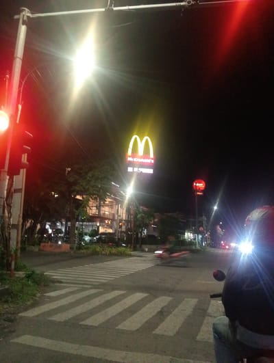A night-time scene unfolds at a traffic intersection in Surabaya, East Java. A bright, illuminated McDonald's sign, with its iconic golden arches, dominates the mid-ground, indicating a fast-food establishment. To the left, a traffic light glows red, and a bright street lamp casts a strong glare, creating lens flare effects across the image. The road features prominent pedestrian crosswalks. In the foreground, partially obscuring the scene, is the back of a person sitting on a motorcycle, their head and shoulders visible, wearing a dark helmet and blue jeans. Their presence suggests the photographer is a road user. The background reveals buildings, trees, and other vehicles, with the headlights of oncoming traffic creating streaks of light. The overall impression is that of a typical urban intersection at night, busy with traffic and illuminated by artificial light. The time of day is clearly night, and the weather appears clear.