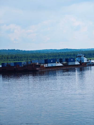A barge loaded with shipping containers is moving along a wide, calm river. The barge is dark red and appears to be made of metal. Several blue shipping containers are stacked on top of the barge. In the distance, a dense forest covers rolling hills. The sky above is partly cloudy with a light blue hue. The water is a deep blue with ripples indicating gentle movement. A white vessel, identified as PT-729 by visible markings, is attached to the rear of the barge, presumably acting as a tugboat. The overall scene suggests a transportation route in a natural, perhaps remote, environment.
