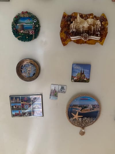 A collection of decorative magnets are affixed to a light-colored wall. From top left, moving clockwise, there is a circular magnet depicting a wreath with bells and a scenic view labeled "ALANYA" with a Turkish flag. To its right is a rectangular magnet made of amber with an embossed image of a castle and figures, labeled "KALININGRAD" and "KÖNIGSBERG". Below this is a square magnet showing a cathedral in "BRNO". At the bottom is a circular magnet enclosed by rope, featuring a beach scene with a starfish and pebbles, labeled "KHAIFA". To the left of the amber magnet is a small, round plate magnet depicting a map of "RHODES". Below that is a rectangular magnet with a grid of nine smaller images, seemingly tourist attractions, with "MINSK" prominently displayed in the center. Between the "Rhodes" magnet and the "Minsk" magnet is a small rectangular magnet showing a building with domes.