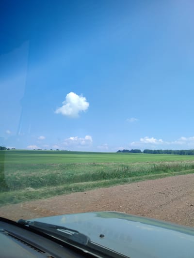 The image depicts a vast, sunlit rural landscape under a clear blue sky with scattered white clouds. In the foreground, the dashboard and windshield of a vehicle are visible, with a wiper blade resting on the windshield. The view through the windshield reveals a wide expanse of green fields, possibly farmland or pasture, stretching towards a distant tree line. A dirt road runs along the right side of the frame, separating the field from the viewer's perspective. There are no people or any signs of urban infrastructure visible in the scene. The overall impression is one of open, natural space.