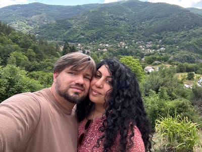 The image is a selfie taken by a couple in a mountainous area near Lukovo, Bulgaria. The man, on the left, has fair skin, light brown hair, and a beard, wearing a brown t-shirt. The woman, on the right, has darker skin, long curly black hair, and is wearing a floral print dress with red sunglasses hanging from her neck. They are positioned in the foreground, with their faces close together. Behind them, the landscape unfolds with rolling green hills covered in dense forest. A village with red-roofed houses is visible nestled in the valley between the hills. The sky is partly cloudy. The overall scene depicts a scenic, rural mountain environment.