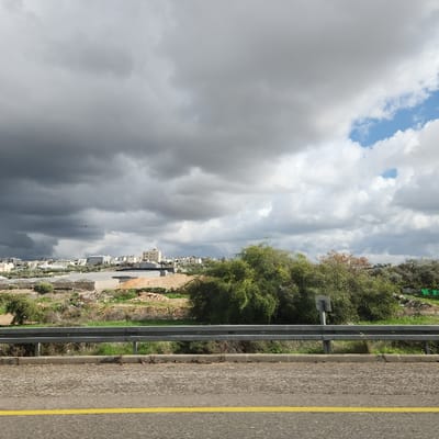 The image displays a landscape within the Menashe Regional Council, Israel, under a sky dominated by heavy, dark grey clouds, with a patch of blue sky and lighter clouds visible in the upper right. The foreground shows a grey asphalt road with a solid double yellow line, bordering a silver metal guardrail. Beyond the guardrail, the midground consists of green vegetation, sections of reddish-brown earth, and several light-colored structures resembling agricultural greenhouses. The background features a dispersed cluster of light-colored, multi-story buildings, consistent with a residential area, interspersed with olive groves. No individuals or discernible activities are present.