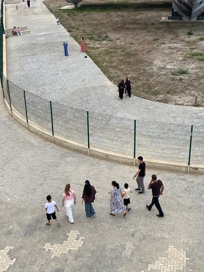 An aerial view captures a paved pathway in a park-like setting, possibly in Arablyar, Russia. A chain-link fence runs along the left side of the pathway, bordering a grassy, dry area. Several individuals are present. In the foreground, a group of three people, a woman in a floral dress, a young boy in a white t-shirt and black shorts, and a man in a dark t-shirt and black pants, are walking away from the camera. Behind them, a man in a black t-shirt and grey pants is walking to the right, and another man in a dark red t-shirt and black pants is walking to the left. Further up the pathway, two women in dark clothing are walking together. There is another woman in a pink top and white pants, and a person in a blue outfit standing and holding a phone. Two women are seated on steps on the left side of the pathway, and a young person is standing in the distance. The ground is covered in a patterned brickwork.