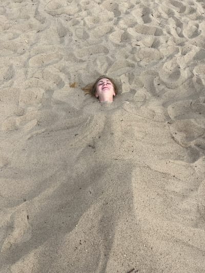 A person's head is visible, sticking out of a mound of sand. They are lying on their back with their eyes closed and a smile on their face. Their hair is blonde and appears slightly damp. The sand is light brown and has been disturbed, with ripples and indentations visible. Some small pieces of debris, possibly twigs or dried seaweed, are scattered across the sand. The lighting suggests it is daytime, possibly a sunny day, as indicated by the shadows cast on the sand. The overall scene conveys a sense of playfulness and relaxation, likely at a beach or sandpit. No visible text is present in the image. The context provided is Загародскі сельскі Савет, Belarus, suggesting a rural location within Belarus.