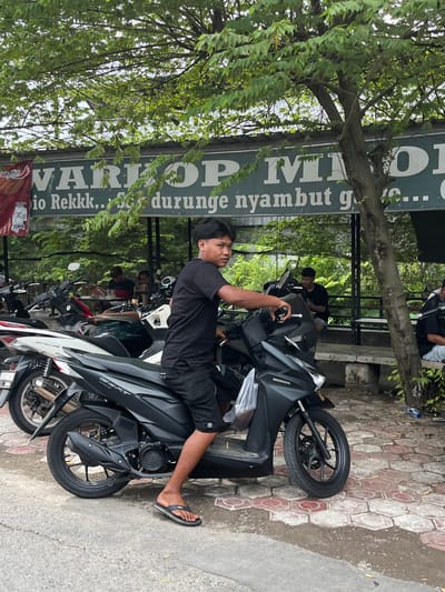 A young man is sitting on a black Honda Beat motorcycle, facing right, in front of a warung (small shop) with green signage. The warung's sign has white text that appears to read "WARJOP MIO" and "io Rekkk.. Sır durunge nyambut g...". The man is wearing a black t-shirt and black shorts, and flip-flops. He is looking slightly to his right. Several other motorcycles are parked around him. In the background, several people are sitting at tables inside the warung, which is surrounded by lush green trees and foliage. The ground is paved with reddish and grey hexagonal tiles. The overall scene suggests an outdoor gathering or resting spot.