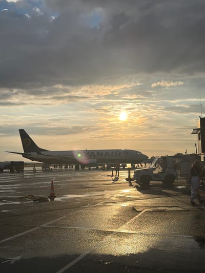 The image depicts an airport tarmac at sunset, with a Ryanair airplane parked and illuminated by the golden light. The tarmac is wet, reflecting the sky and the setting sun, creating a shimmering effect. Several people are visible on the tarmac, some walking and others standing near the aircraft. Airport vehicles, including a baggage tug, are also present. The sky is filled with a mix of dramatic, dark clouds and brighter, illuminated patches where the sun is breaking through, casting long shadows. The overall atmosphere is serene and evocative of the end of a day at the airport. The location is identified as the city of Tillé, France.