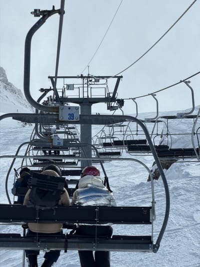 The image depicts a view from a ski lift, likely in La Giettaz, France, given the snowy mountain environment. The camera is positioned looking down the lift line, showing several chairs. Two people are seated in the foreground chair. The chairlift structures and cables are visible, along with the snow-covered terrain below. Chair numbers 38, 39, 36, and 7 are visible. The sky is overcast, creating a muted light.