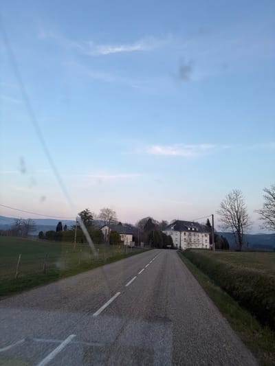 This image depicts a rural road leading into a settlement within Le Saulcy, France, under conditions suggesting twilight. The asphalt road, running from the foreground towards the background, is marked with a continuous white line on its left edge and a dashed white center line.
On the left side of the road, a grassy field is segmented by a fence composed of wooden posts and wire. Beyond this field, a cluster of deciduous trees in a bare state for the season, and at least two structures, one a white building with a dark roof, are visible amidst further foliage. Rolling hills are visible on the distant horizon.
On the right side of the road, a grassy verge separates the roadway from a large, open field. Further down this side, a multi-story white building with a dark, complex pitched roof stands prominently. This building has multiple windows and appears to be a substantial structure. To its immediate right, a road sign with a black arrow on a white background points left, indicating a directional change in the road. Bare deciduous trees are present both in the foreground and surrounding the building. A utility pole stands near the white building.
The sky is predominantly light blue, transitioning to a pale pink-purple hue at the horizon. Wispy white clouds are scattered across the upper sky. A prominent diagonal streak of light glare traverses the image from the upper left to the lower right, and several circular lens flares are also present. No individuals or vehicles are visible in the scene.