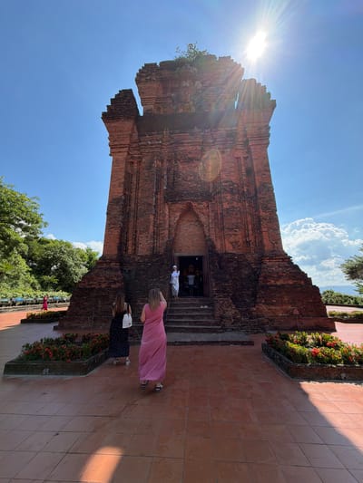 A wide-angle, low-angle shot captures the Po Klong Garai Cham Towers in Tuy Hoa Ward, Vietnam. The ancient brick structure dominates the frame, its weathered red bricks showing signs of age and deterioration, particularly at the top. The sun shines intensely from the upper right, creating lens flare and casting a bright glow that obscures some details of the tower.

Two women stand in the foreground, facing away from the camera, looking towards the tower entrance. The woman on the left wears a black and white polka dot dress and carries a white tote bag. The woman on the right is dressed in a long, flowing pink dress. Both are standing on a reddish-brown tiled plaza.

Several people are visible entering the tower through a dark arched doorway, which is accessed by a set of stone steps. A figure in white is standing at the top of the steps.

Flanking the plaza are beds of vibrant green foliage dotted with small red flowers, adding a splash of color to the scene. Lush green trees and vegetation are visible on the left side of the image, and a large, fluffy white cloud drifts in the bright blue sky on the right. The surrounding landscape is hilly and appears somewhat arid.

The overall atmosphere is one of historical grandeur bathed in bright sunlight.
