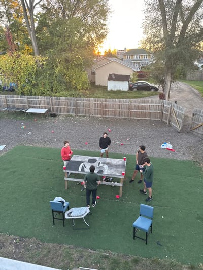 This image captures an aerial view of an outdoor gathering in a residential backyard in South Bend, United States, at dusk or late afternoon. The scene features five young adult males engaged in an activity around a large, weathered wooden table on an artificial turf area.
The primary activity center is the large wooden table, painted with a white "69" or similar design, typical of a beer pong setup. Numerous red plastic cups are placed on and around the table, with many more scattered across the artificial turf and a gravel area to the left and behind the table. Two light blue high-backed chairs and a small white side table are positioned on the artificial turf to the left of the main table.
The five males are positioned around the large table. One male in a red shirt stands at the left end, holding a red cup. Another in a black hoodie and light shorts stands at the opposite end. Two males in dark green and brown shirts/hoodies are on the right side of the table; both appear to be holding small, dark objects, possibly game pieces or balls. A fifth male in a dark green shirt stands with his back to the viewer on the near side of the table.
Beyond the artificial turf and activity area, a gravel and dirt expanse is visible, littered with additional red cups and miscellaneous debris. This area also contains two smaller white folding tables. A wooden fence separates this immediate backyard from adjacent properties. To the left, dense green foliage and residential structures are visible. To the right, the wooden fence has a partially open gate leading to a dirt path. A black sedan is parked near a small white shed in a grassy area behind the fence, with other residential houses and trees in the background. The sun is low in the sky, casting a warm, golden light on the distant trees and structures.