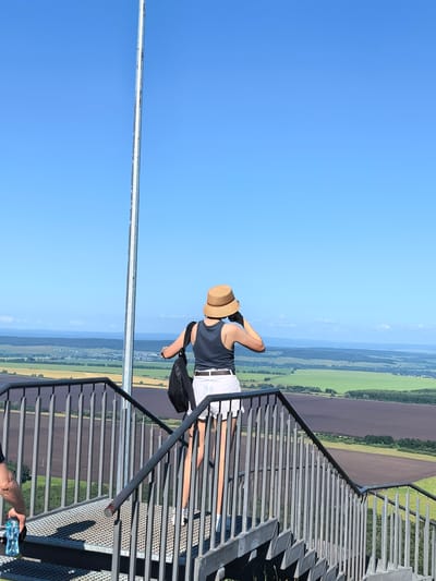A person wearing a straw hat, a dark tank top, and white shorts stands on a metal staircase, looking out at a vast landscape of fields and hills under a clear blue sky. The person is holding a black bag over their left shoulder and appears to be taking a photo with their right hand, holding a device up to their face. The staircase has a metal railing and a grated metal surface. In the foreground, another person's arm is visible holding a blue water bottle. The background reveals a panoramic view of agricultural fields in various shades of green and brown, with a small town or village nestled in the distance. The overall scene suggests a viewpoint or observation deck in a rural or semi-rural area, possibly in Shikhan, Russia, given the context.