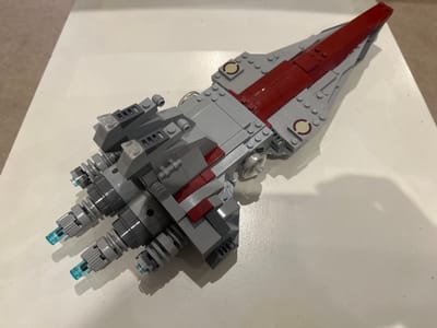 A static, elevated perspective captures a LEGO brick model of a spacecraft, approximately 30-40 cm in length, resting on a flat, off-white surface. The model is predominantly grey with a dark red central spine running along its dorsal length. The stern features four primary engine thrusters, each containing a translucent blue element simulating exhaust, flanked by smaller grey thruster units and structural components. Additional circular light tan elements with dark red rings are integrated into the grey sections of the hull. Two smaller grey structures topped with translucent clear rectangular pieces are positioned dorsally towards the rear. The model casts a distinct shadow on the surface, indicating overhead illumination. A textured, light brown surface is partially visible in the upper-left background. No individuals or active interactions are depicted. This scene is located in the city of Abbotsbury, United Kingdom.