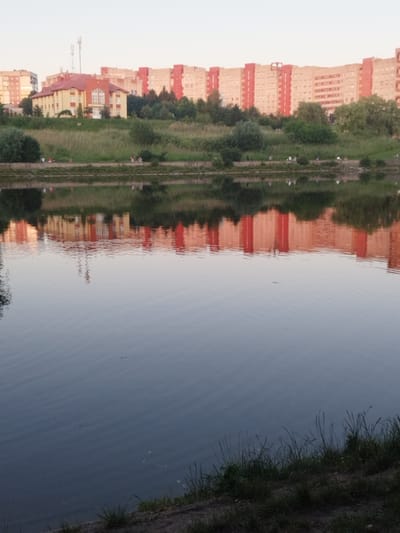 The image depicts a waterfront scene in Novyi Rozdil, Ukraine, likely during the late afternoon or early evening due to the soft lighting. In the background, a row of tall, multi-story apartment buildings with red and beige facades stretches across the horizon, interspersed with trees and greenery. A smaller, yellow building with a red roof is prominent in the left-center of the background. A body of water, possibly a river or lake, occupies the foreground, reflecting the buildings and sky. Along the grassy bank of the water, several individuals are visible sitting and interacting, suggesting a recreational area. The water's surface is rippled, distorting the reflections. The overall atmosphere is serene, with a mix of urban development and natural elements.