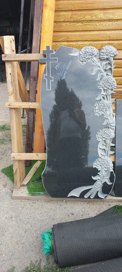In Ertis, Kazakhstan, a black granite headstone stands propped against a wooden structure. The headstone is intricately carved with a cross at the top left and a floral arrangement along the right edge, featuring several blooms and a ribbon. Inscribed on the stone are the numbers "390T", the name "Иван С.", and the text "Размер 120x60". A reflection of a person wearing a dark t-shirt is visible on the polished surface of the headstone. The scene is outdoors, with a concrete ground, some grass, and a rolled-up mat visible in the foreground. The background consists of wooden planks and metal bars, suggesting a workshop or storage area.
