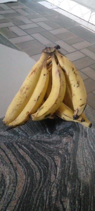 A cluster of approximately 7-9 ripe, yellow bananas with scattered brown blemishes rests on a dark, horizontally striated countertop. The countertop exhibits wavy patterns of black, grey, and reddish-brown, indicative of granite or similar stone. The background features a lighter-toned floor or wall composed of rectangular tiles arranged in a herringbone pattern. No individuals or active interactions are present. The scene is situated indoors, in Agunmo, Nigeria.