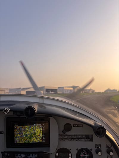 The image captures a first-person perspective from the cockpit of a light aircraft at Toussus-le-Noble Airport (LFPN), France, during a low sun angle period (likely sunrise or sunset).

In the foreground, the top section of a grey and white aircraft dashboard is visible. Embedded in the dashboard is a digital navigation screen displaying a detailed, multi-colored geographical map, indicative of an urban or semi-urban area with roads, waterways, and possibly airspace information. The screen displays a timestamp of "17:53" and menu options like "Zoom Out," "Direct To," and "Return." Below this screen, a portion of radio frequency displays shows "120.750" and "123.500." To the right of the navigation screen, a rectangular plaque with text is affixed to the dashboard, reading in part: "R100 AIRPLANE. APPROVED IN ACCORDANCE FOR VFR DAY/NIGHT OPERATIONS. FLIGHTS IN KNOWN ICING CONDITIONS PROHIBITED. AEROBATIC MANOEUVRES INCLUDING SPINS PROHIBITED. REFER LIMITATIONS ACC. TO AIRPLANE FLIGHT MANUAL." Other cockpit elements include air vents, knobs labeled "NAV INSTR" and "ENGINE INSTR," and a "ALIM. 12V DC" input.

Through the aircraft's windshield, the exterior environment is visible. The sky transitions from a pale blue in the upper left to a warm orange-yellow hue towards the right, suggesting either early morning or late afternoon. In the mid-ground, multiple hangars or airport buildings are present, slightly out of focus. To the left, the distinct T-tail empennage of another parked aircraft is partially visible. To the right, blurred propeller blades indicate an aircraft that is either taxiing or has its engine running. A paved taxiway or runway surface is visible leading into the distance on the right, bordered by green grass. No individuals are explicitly visible, but the active cockpit and surrounding aircraft suggest ongoing airport operations.