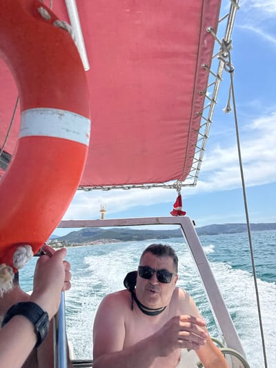 In this medium shot, a man wearing sunglasses and a black choker is onboard a boat. He is shirtless and appears to be speaking to the camera. His right hand is raised, palm facing outward, in a gesture of speech or emphasis. Beside him, a person wearing a black smartwatch is holding onto a metal railing. In the foreground, a bright orange life preserver with a white stripe is prominently displayed, attached by ropes to the boat's railing.
The scene is set on a body of water, likely the sea, with waves and white spray indicating the boat is in motion. In the background, a coastline with hills and buildings is visible under a bright, partly cloudy sky. The sun is high in the sky, suggesting it is daytime, possibly midday. The overall mood is casual and adventurous, suggesting a leisure activity like a boat trip. The presence of the life preserver emphasizes a maritime setting and the potential for activities on or near the water. No visible text is present in the image. The location context provided is Limenaria, Greece.