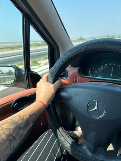 This is a first-person perspective shot from inside a car, looking towards the dashboard and steering wheel. The subject is a person's left hand, with a floral tattoo visible on their forearm and a thin red string bracelet on their wrist, holding the steering wheel. The steering wheel is black with a quilted texture on the outer rim and features a Mercedes-Benz logo in the center, along with "SRS AIRBAG" text.
The dashboard displays a speedometer, with the needle indicating a speed of around 20 km/h. Part of the car's interior, including a brown trim piece and an air vent, is visible. Through the windshield and side window, a bright, clear sky suggests it is daytime with good weather. The view outside shows a highway with multiple lanes bordered by dry, sparse vegetation.
The overall impression is one of a casual drive on a highway, possibly in a sunny, rural or suburban area. The focus is on the car's interior and the driver's immediate surroundings, giving a sense of being in motion. The visible text on the dashboard is primarily related to the car's instrumentation and safety features.