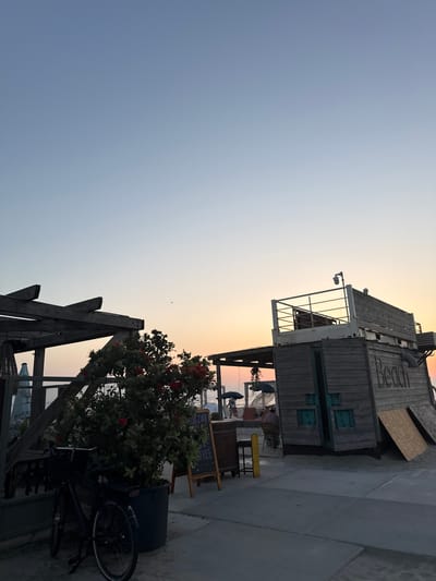 The image depicts a beachside scene in Hook of Holland, Netherlands, at dusk. The sky is a gradient of pale blue to soft orange near the horizon. In the foreground, a dark-colored bicycle is parked next to a large black planter holding a lush green bush with red flowers. To the left, a wooden structure with exposed beams and a woven hanging basket is visible. To the right, a rustic wooden building with the word "beach" painted on its side stands prominently. This building has an upper deck with a railing and a security camera. A small awning extends from the building, casting a shadow. Further in the background, beach umbrellas and a few distant figures can be seen. A chalkboard sign with some text, possibly indicating "FREE," is propped up near the planter. The ground is a mix of sand and paved surfaces. The overall atmosphere suggests a relaxed, end-of-day setting by the sea.