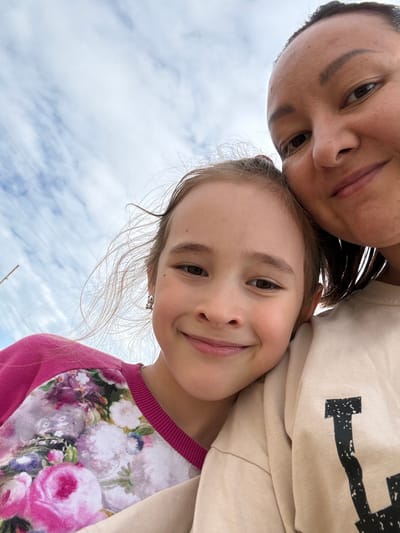This is a low-angle, close-up selfie taken outdoors, featuring two individuals, likely a mother and daughter, against a backdrop of a cloudy sky. The woman, on the left, has dark hair and is looking down towards the camera. Her facial features are visible, including her eyes, nose, and mouth. The girl, positioned slightly in front of her, is also looking at the camera and smiling broadly, revealing her teeth. She has light brown hair, and a small earring is visible on her left ear. Her attire consists of a pink long-sleeved shirt with a floral pattern featuring pink and white roses. The woman appears to be wearing a light-colored beige sweatshirt with a black graphic design on it. The photo captures a moment of shared joy and connection between the two. The lighting suggests it's daytime, possibly late afternoon given the soft light, and the cloudy sky indicates moderate weather conditions. There are no other significant environmental cues to pinpoint the exact location, other than the provided context of Большой Турыш, Russia.